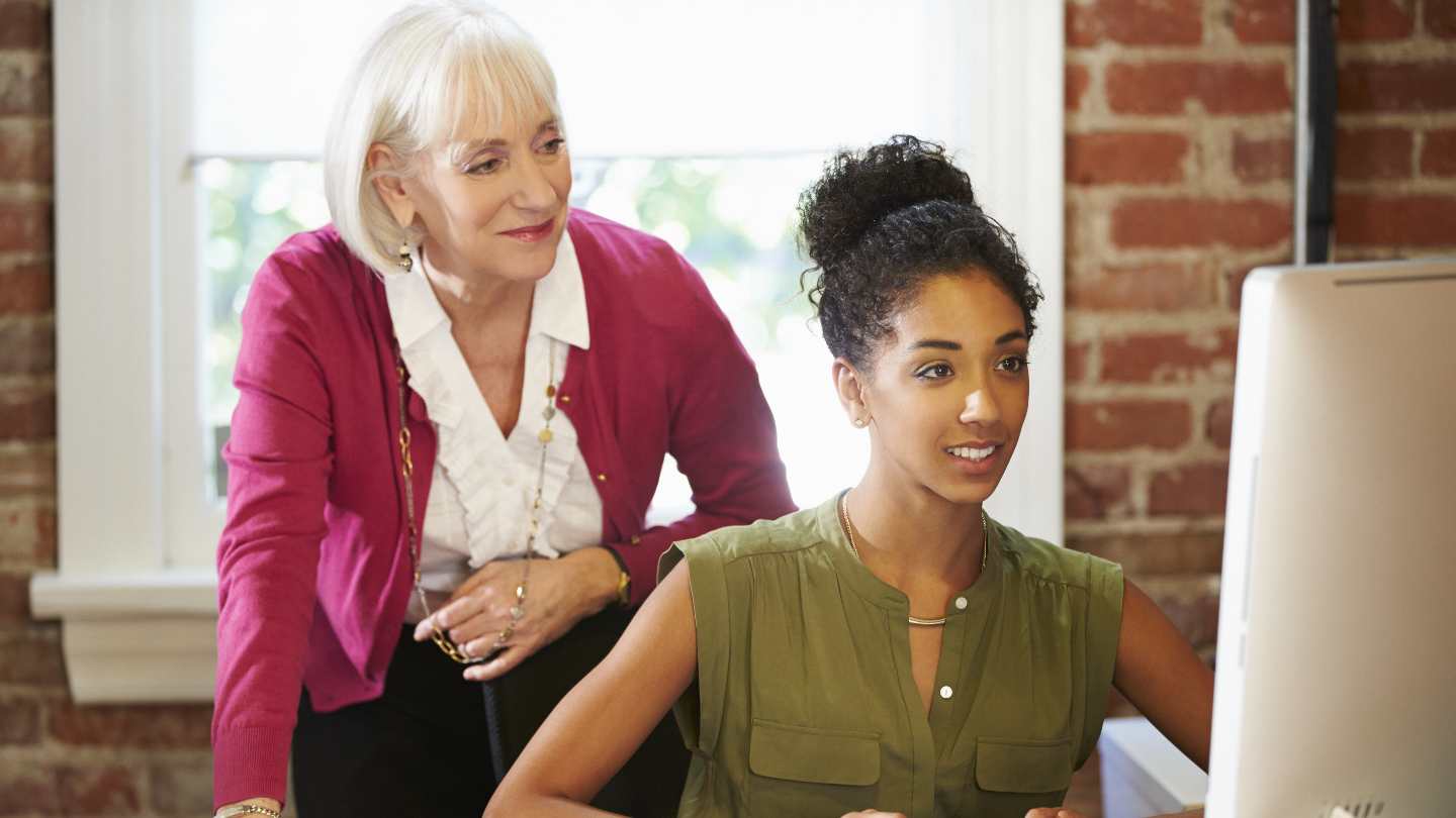 15-ways-young-tech-pros-can-find-a-perfect-mentor-match Mentor over employee's shoulder while she is on the computer.