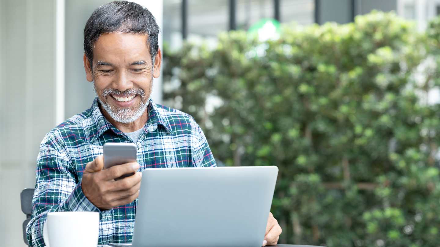 apps-to-help-leaders-reach-their-new-years-resolution Man smiling at phone while his laptop is open outside at a cafe.